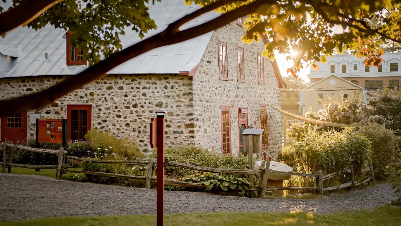 Maison ancestrale en pierre d'un village du Qu&eacute;bec, entour&eacute;e d'arbres aux couleurs automnales et d'un sentier bord&eacute; d'un lampadaire au coucher du soleil